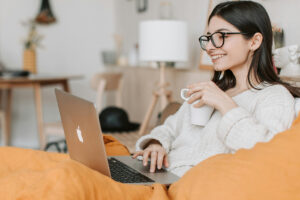 Kvinde sidder i sofa med kaffekop i hånden og smiler, mens hun kigger på sin bærbare computer.